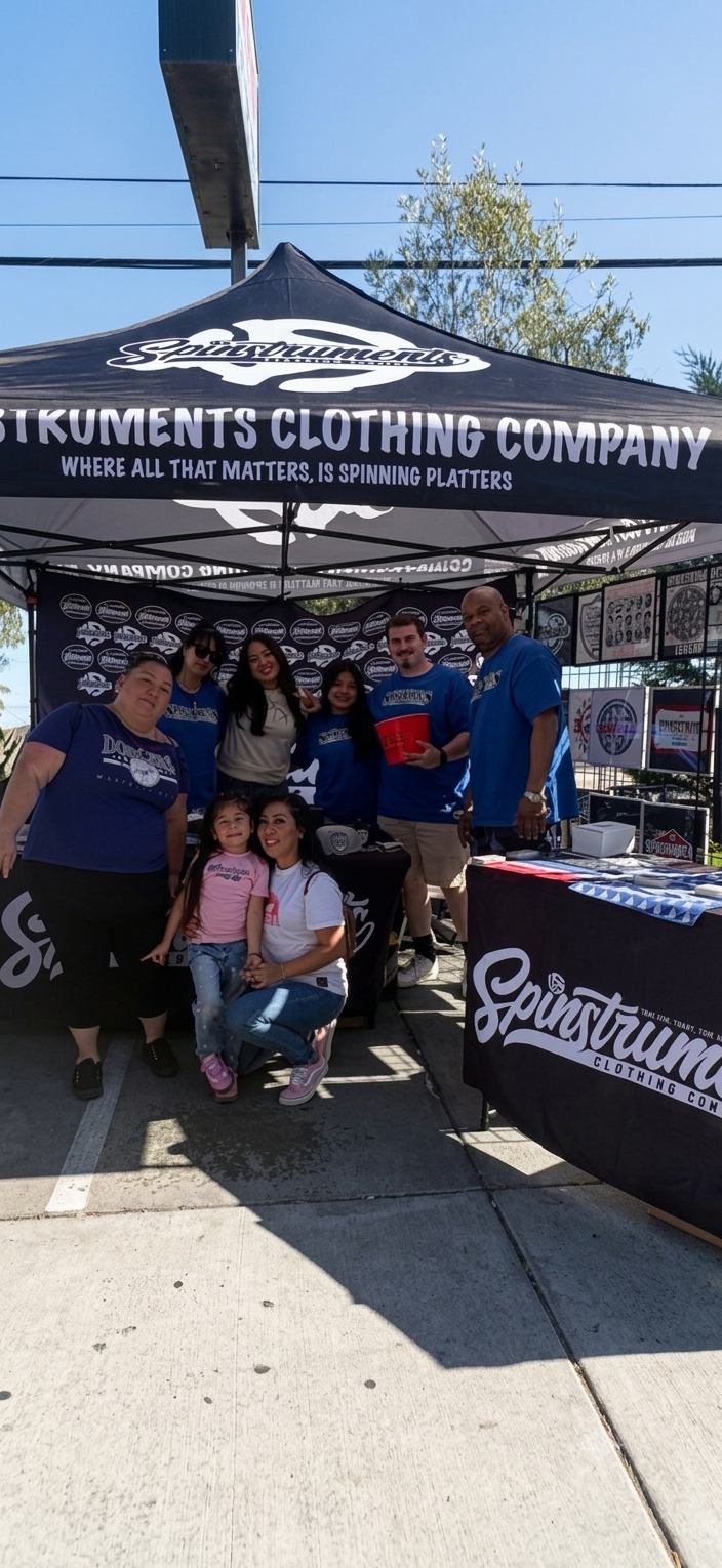 A group of people posing under a black pop-up canopy tent branded 'Gnarsstrum Instruments Clothing Company' outdoors on a sunny day, with a sidewalk and trees in the background.
