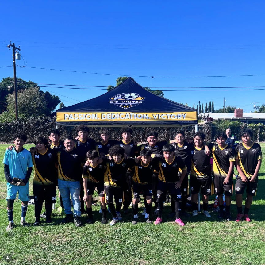 A group of young soccer players posing outdoors on a grassy field in front of a black and yellow canopy tent. Several players are wearing black jerseys with yellow stripes and matching black shorts, while one player on the left wears a light blue jersey and another wears jeans.