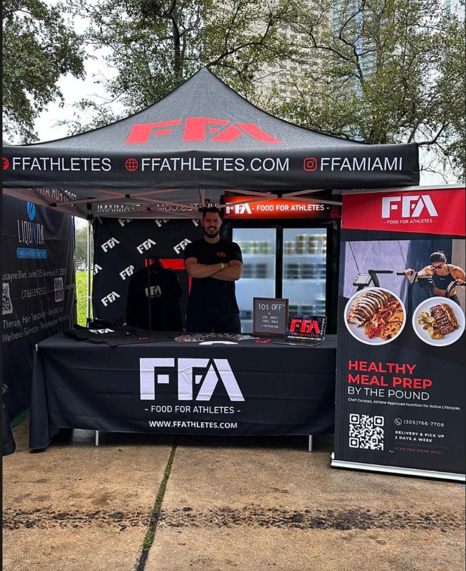 An outdoor promotional booth for FF Athletes / Food For Athletes under a black canopy tent with red and white branding. A man stands behind the table, and printed banners and a sign are displayed on the front and to the right.