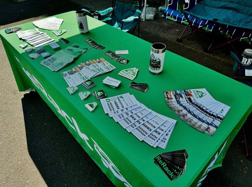 A green promotional table covered with printed materials, stickers, cards, and branded cups at an outdoor event. The tablecloth and printed items prominently show the ReRack brand name.
