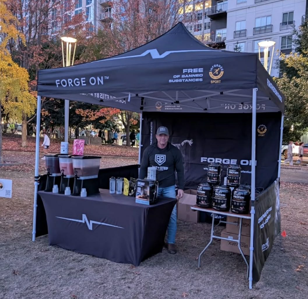 A black pop-up canopy tent branded FORGE ON is set up outdoors in a park-like area with trees and buildings in the background. A man stands behind the tent at a display table with drink containers and supplement cans.