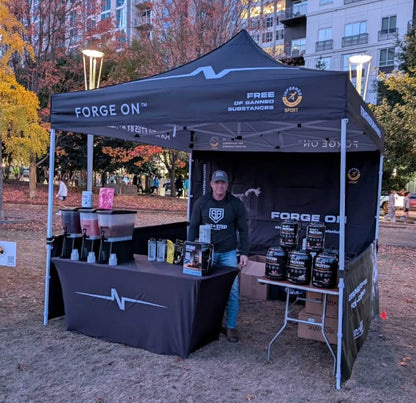 A black branded pop-up canopy tent set up outdoors in a park-like area, with a person standing behind a table inside the tent. The canopy and tablecloth show the brand name Forge On, along with supplement containers displayed on the table and inside the tent.