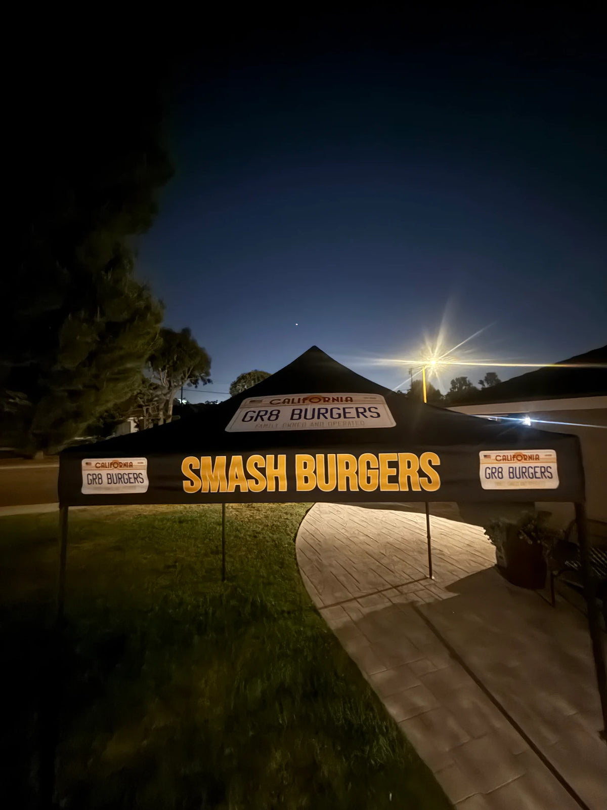 A black event canopy tent with 'SMASH BURGERS' in large yellow letters, branded with 'California GRB BURGERS' on the top and side panels, set up at night on a grassy lawn next to a paved walkway.